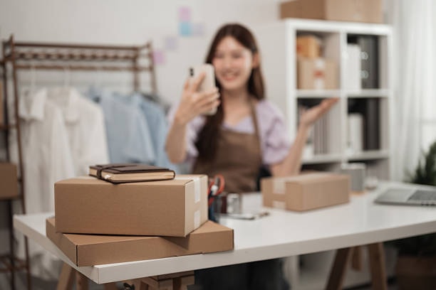 E-commerce seller using smartphone with cardboard shipping boxes and packing supplies on desk