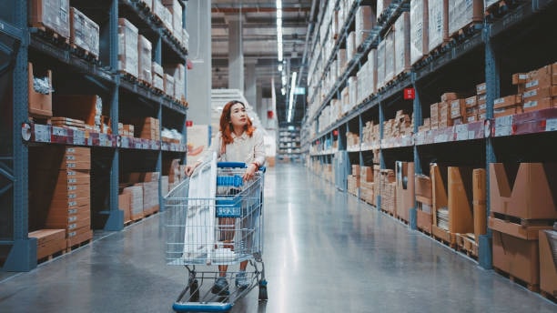 Woman pushing shopping cart in large warehouse aisle with shelves full of packaging supplies