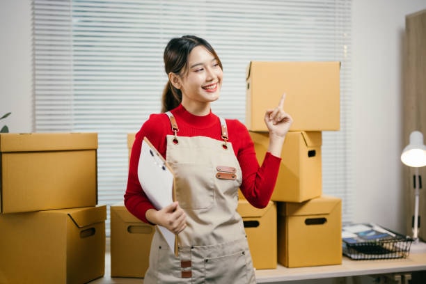 Young woman small business owner in red shirt standing with brown cardboard boxes in workspace