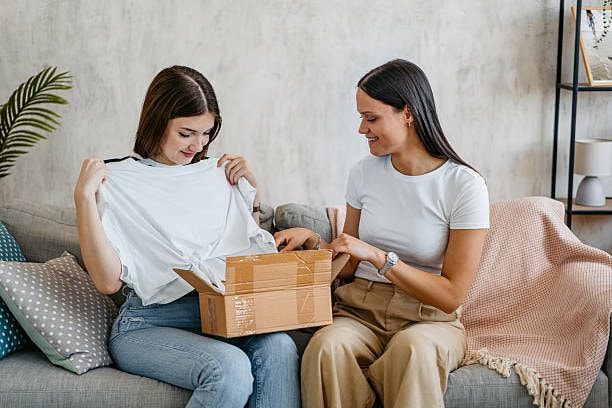 Two women sitting on couch opening a cardboard box and revealing a white T shirt