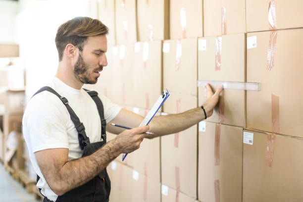 Man in overalls using a ruler to measure boxes stacked in a warehouse