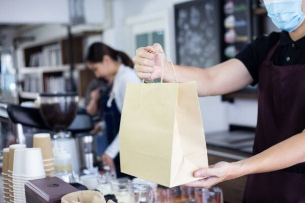 Barista wearing an apron and mask holding a kraft takeout paper bag at a coffee shop counter.