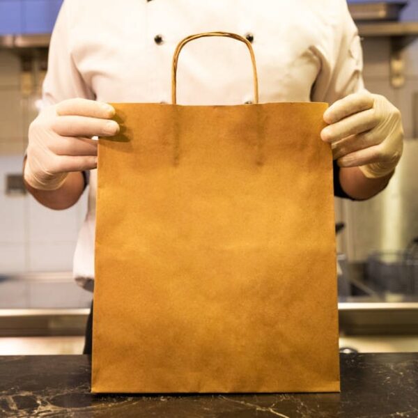 Chef in uniform holding a large brown paper takeout bag with handles in a kitchen.