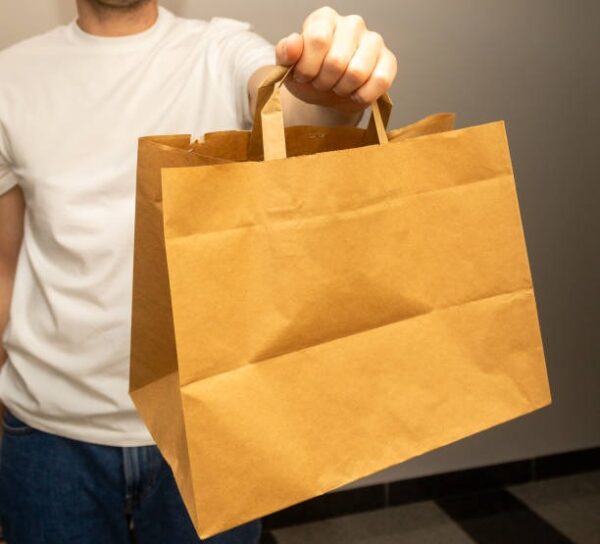 Person in casual clothes presenting a brown paper takeout bag toward the camera.