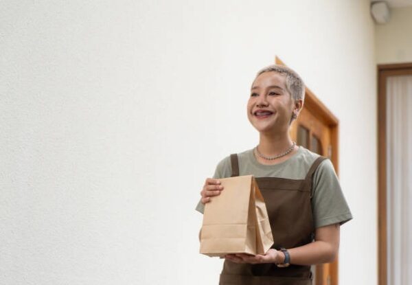 Female restaurant staff smiling while holding a small takeout paper bag for delivery.
