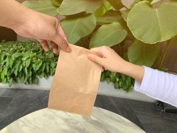 Close-up of two hands exchanging a small kraft takeout paper bag in an outdoor setting.