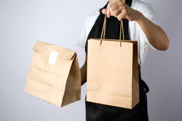 Worker wearing apron holding a folded paper lunch bag and a handled takeout paper bag.