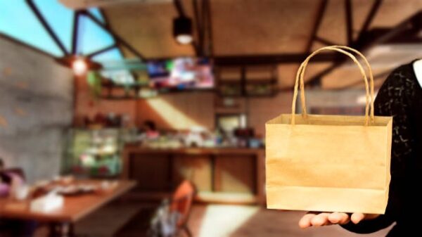 Small kraft paper bag with handles placed on a customer’s hand inside a café.