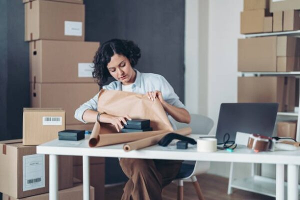 Woman sitting at a desk wrapping products with kraft paper surrounded by cardboard boxes.