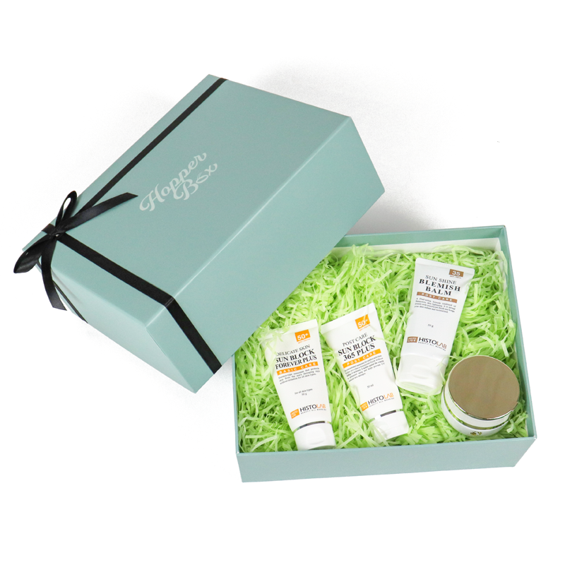 Light blue rigid gift box with black ribbon and branded lid, containing skincare products like sunscreen and blemish balm nestled in green shredded paper fill—ideal for custom cosmetic packaging that balances protection and presentation.