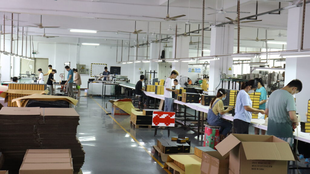 Factory floor with workers assembling rigid boxes and folding cartons, stacks of cardboard, and production lines, showcasing high-volume packaging manufacturing.