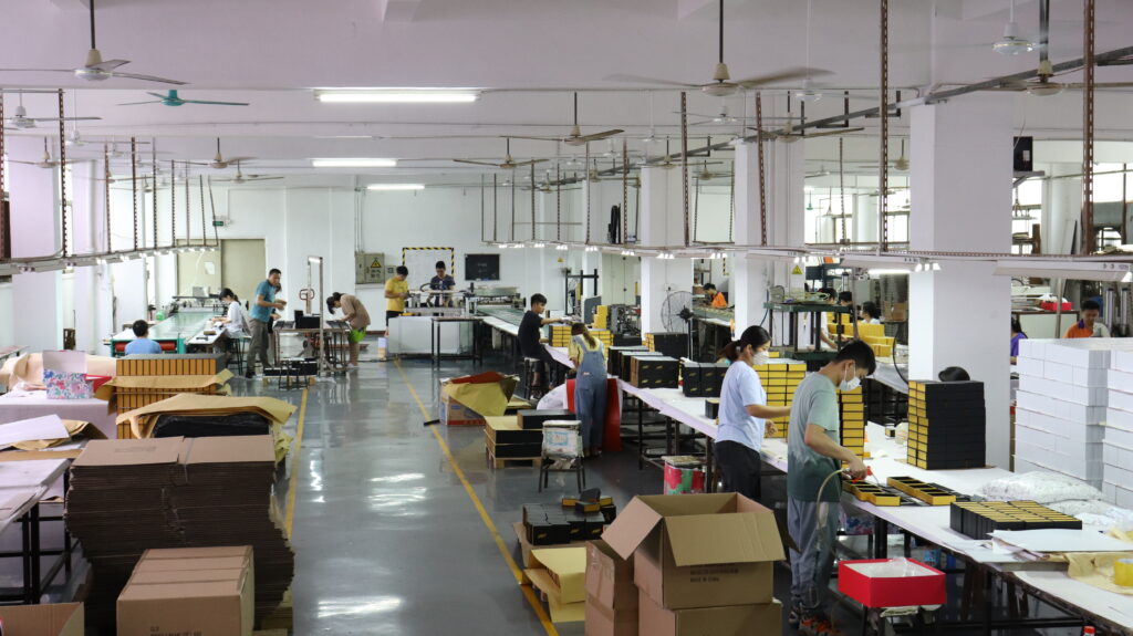 Wide-angle view of a packaging factory floor with workers assembling die-cut boxes at long tables, surrounded by stacks of finished products and raw materials.