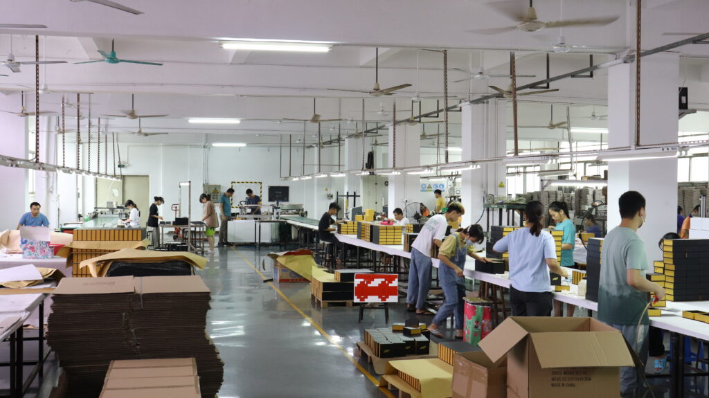 Inside a packaging factory, workers operate industrial printing and die-cutting machines while assembling rigid boxes and folding cartons—demonstrating the production process behind high-volume custom apparel packaging solutions tailored for both e-commerce and retail channels.