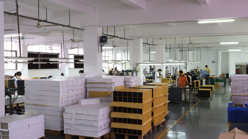 Interior view of a packaging factory with stacks of finished boxes and workers assembling custom rigid boxes, highlighting the industrial scale of blind and foil embossing production.