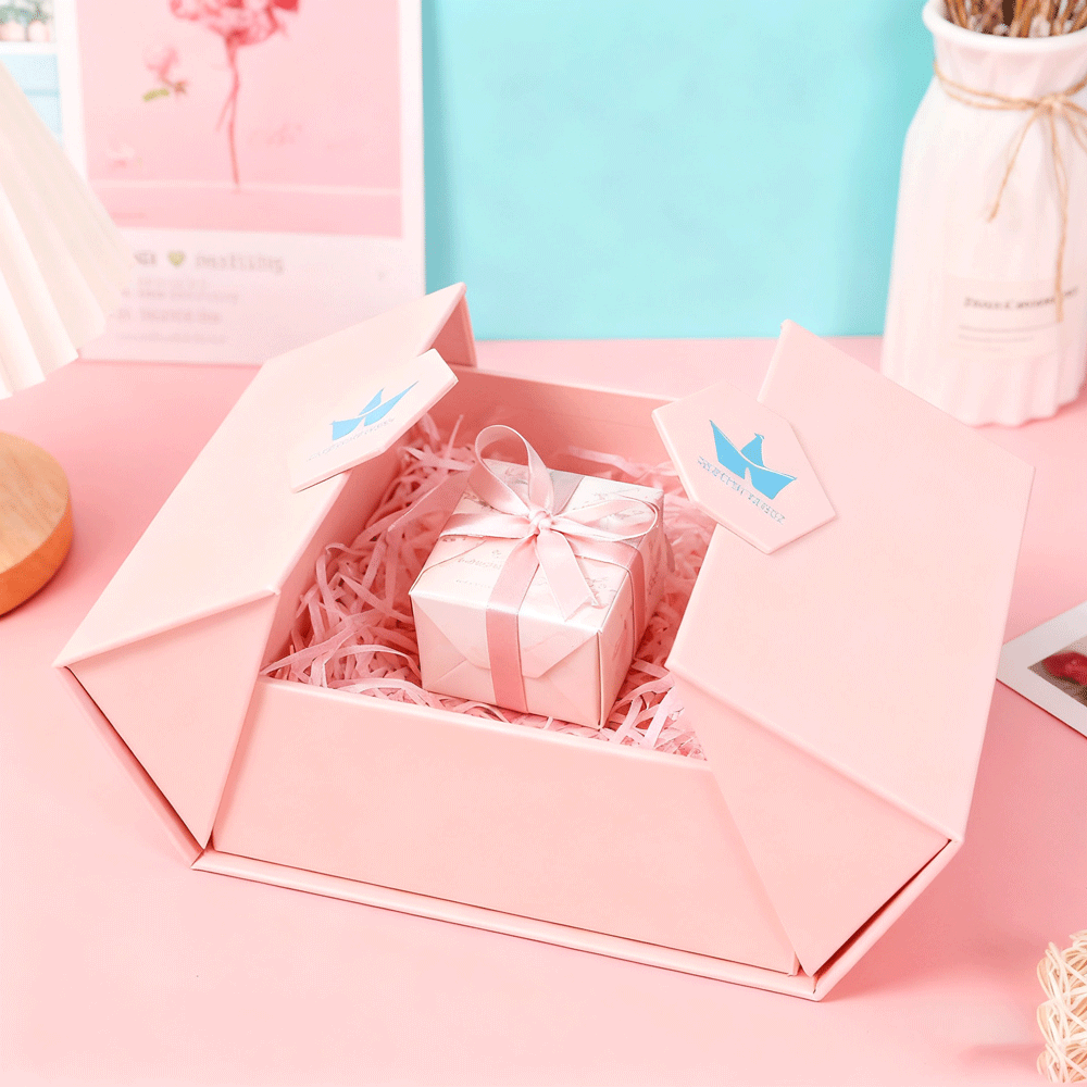 Open pink gift box with shredded paper filler and a small wrapped present inside, featuring a matching logo on flaps—ideal for custom packaging using precise color matching via Pantone or CMYK.