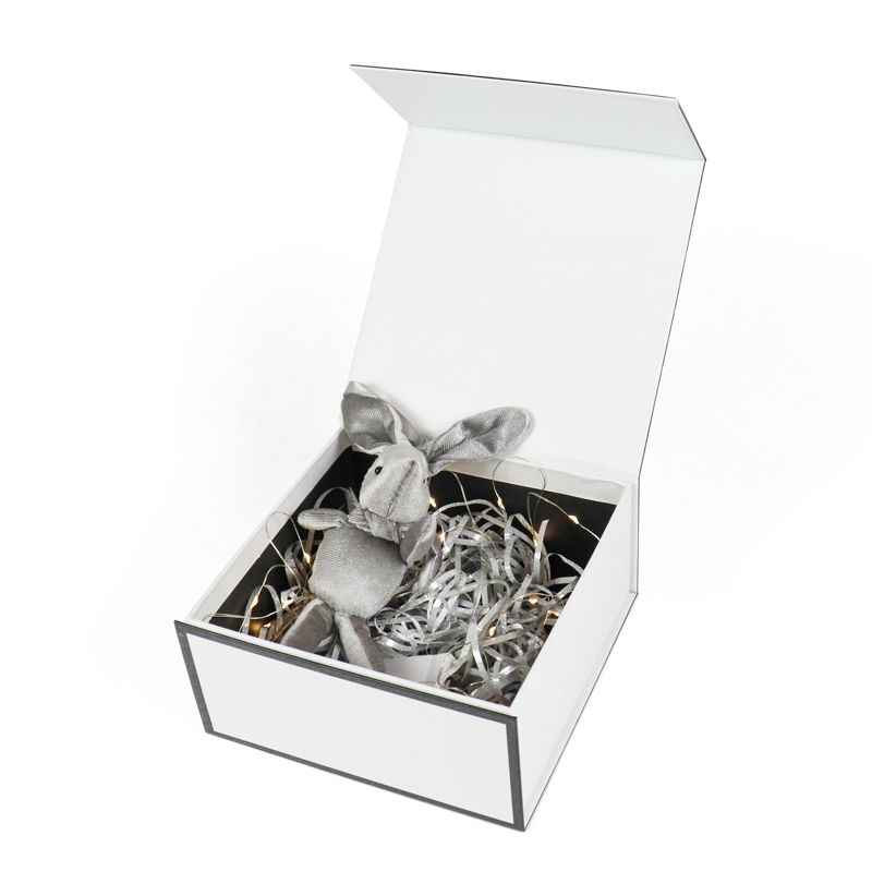 A white gift box opened to reveal a silver plush bunny nestled among shimmering shredded paper and delicate fairy lights—creating a magical, emotionally engaging unboxing experience tailored for special occasions or limited-edition releases.