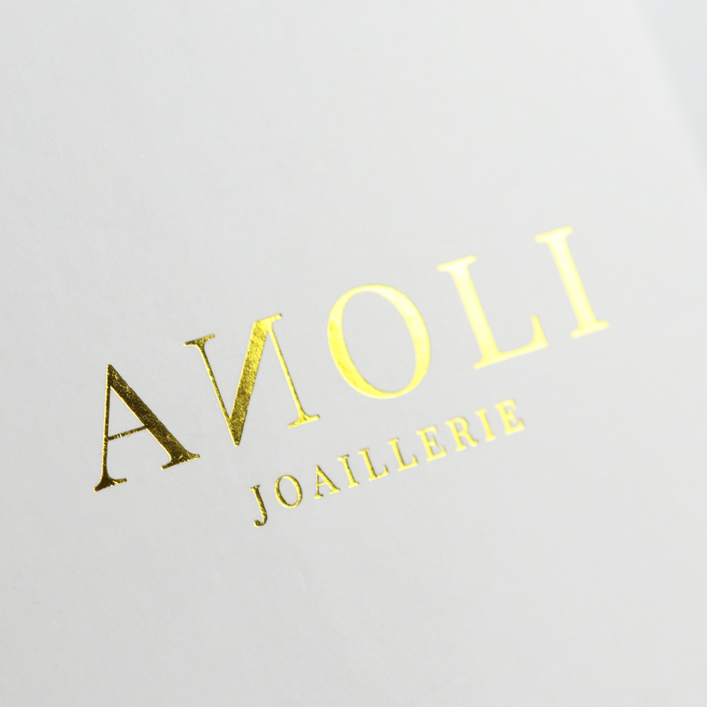 Close-up of gold foil-stamped logo "ANOLI JOAILLERIE" on a white matte surface, showcasing high-gloss metallic finish and sharp registration typical of hot stamping for luxury jewelry packaging.