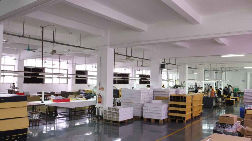 Wide-angle view of a packaging factory floor with workers assembling boxes and stacks of finished products ready for shipping.