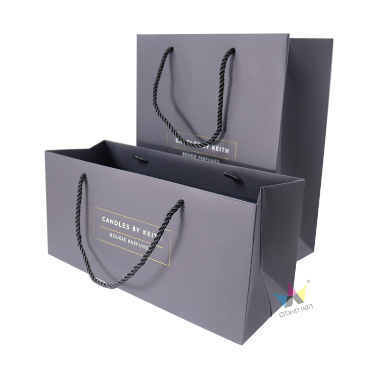 Elegant dark gray paper gift bags with twisted rope handles and gold foil branding, designed for luxury product presentation such as candles, accessories, or premium apparel sets.