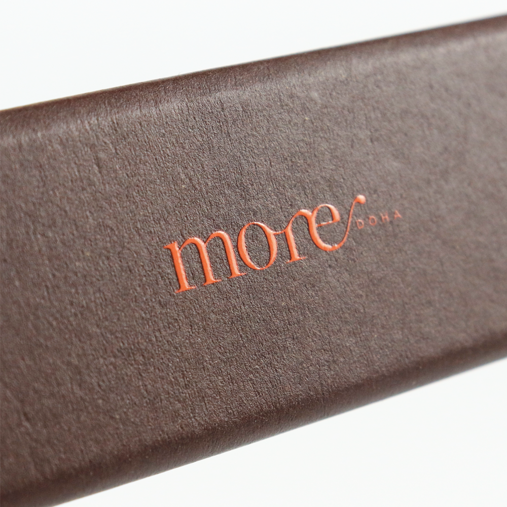 Close-up of a brown textured box with an orange "more DOHA" logo featuring a glossy Spot UV coating, creating a high-contrast visual effect against the matte background—ideal for premium brand identity.