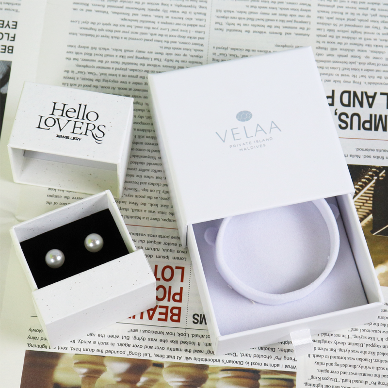 Open book-style jewelry box with printed text cover, revealing two pearl stud earrings securely placed in a black velvet-lined compartment, showcasing elegant and precise custom packaging.