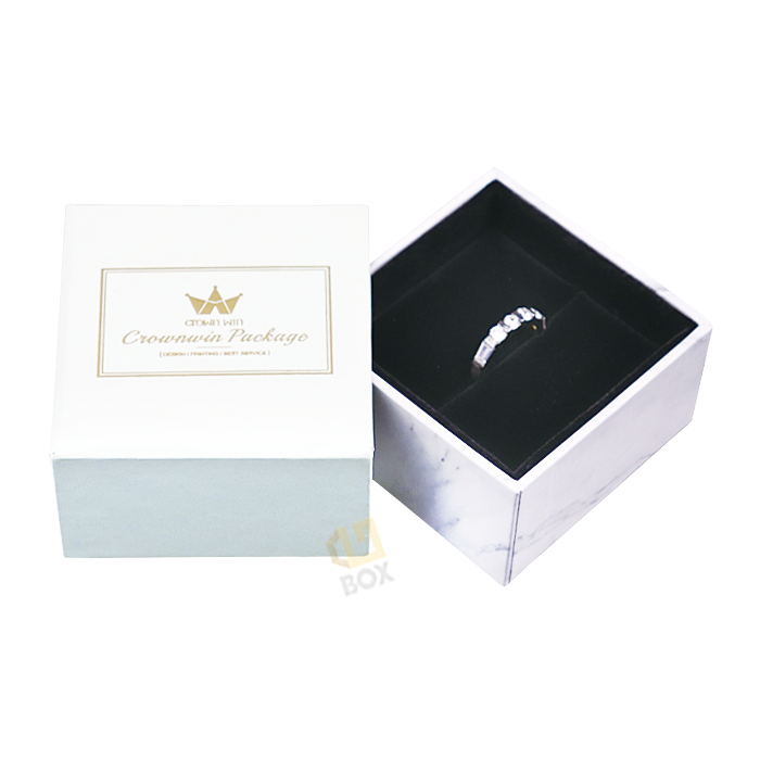 Open white rigid jewelry box with marble-patterned sides and gold foil logo, revealing a diamond ring securely placed in a black velvet-lined interior, showcasing precision insert design.