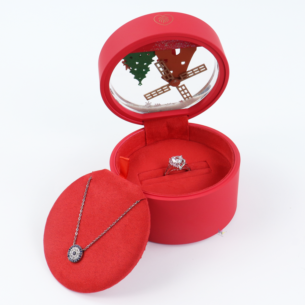 Open red round jewelry box with transparent lid showing a Christmas-themed snow globe scene, featuring a diamond ring securely placed in a custom insert and a separate velvet tray holding a pendant necklace.
