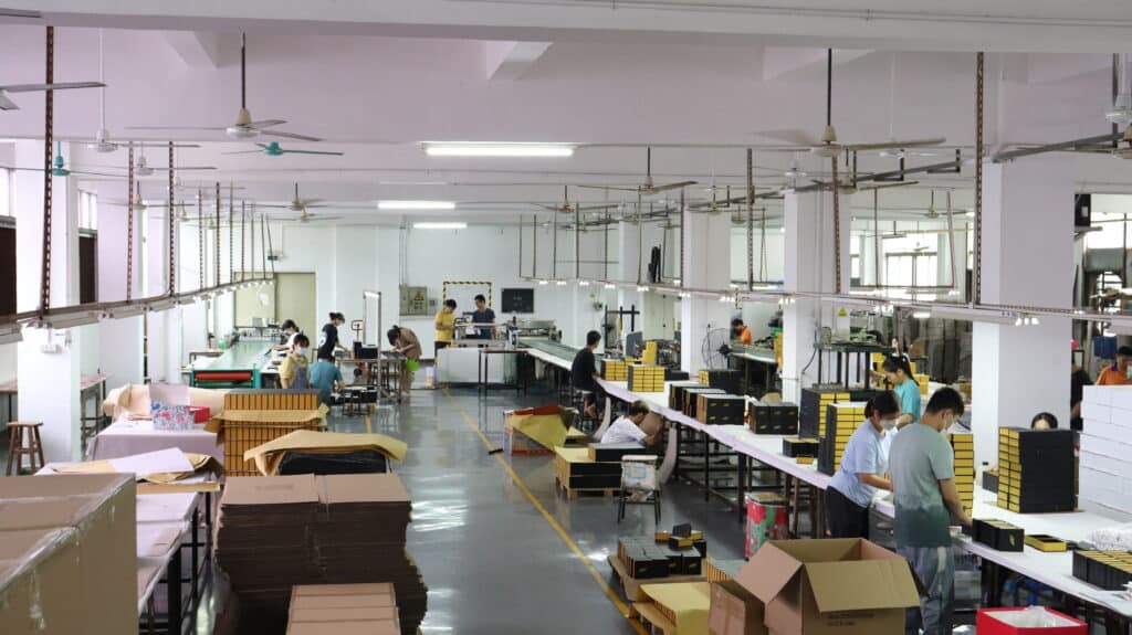 Wide-angle view of a busy packaging factory floor with workers assembling wine boxes, stacks of materials, and production lines for rigid boxes and inserts.