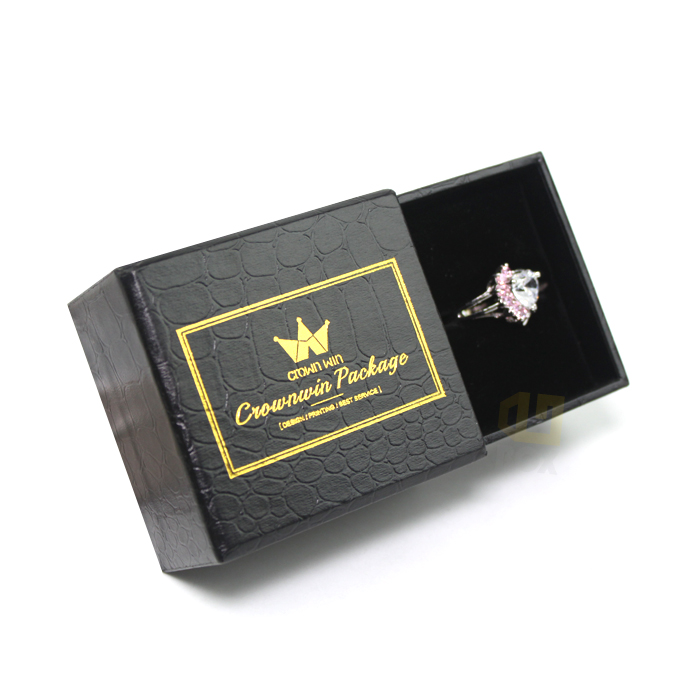 Black rigid jewelry box with crocodile-embossed surface and gold foil-stamped logo, open to reveal a diamond ring securely held in a velvet-lined interior, showcasing high-end custom packaging design.
