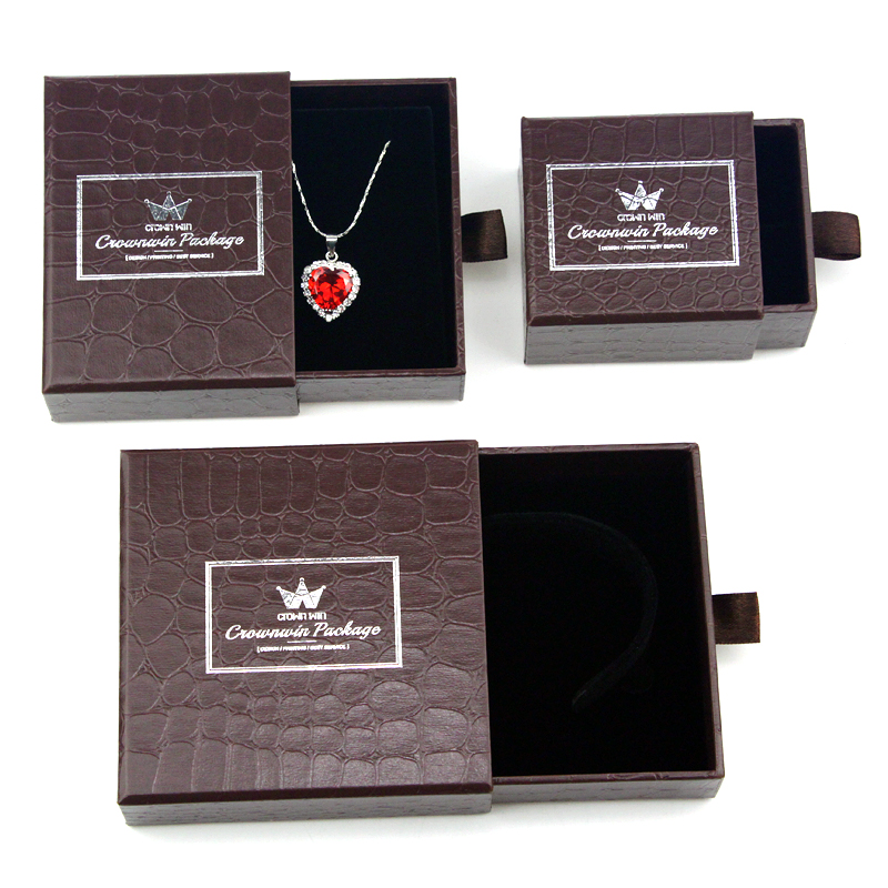 Three dark brown crocodile-textured jewelry boxes with white foil-stamped logos, one open with a heart-shaped pendant necklace, another with a headband, and one closed, showcasing custom packaging design and structural integrity.