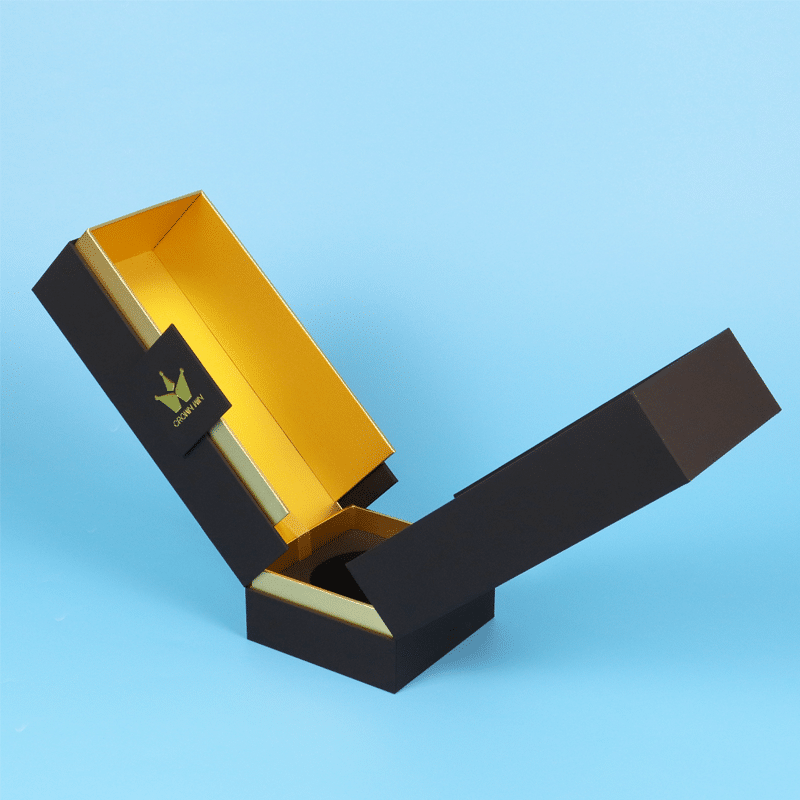 Stylish black and gold rigid wine box with angular, sculptural design featuring a hinged lid and golden interior, displayed on a white textured surface against a blue background.