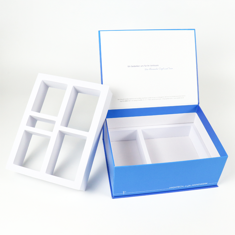 Open blue rigid box with white paperboard insert featuring geometric compartments; lid displays handwritten-style message in German, "Wir bedanken uns für Ihr Vertrauen."