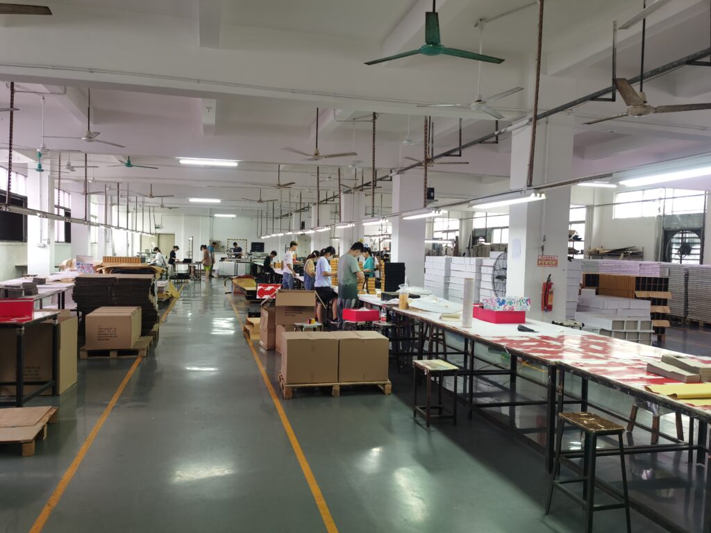 Wide view of a packaging factory floor with workers assembling boxes, stacks of materials, and long worktables under industrial lighting.