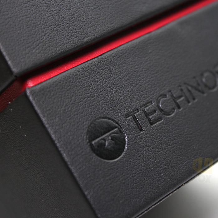 Close-up of black leather-textured box with debossed "TECHNO" logo and red accent line, showing fine edge detail and texture.