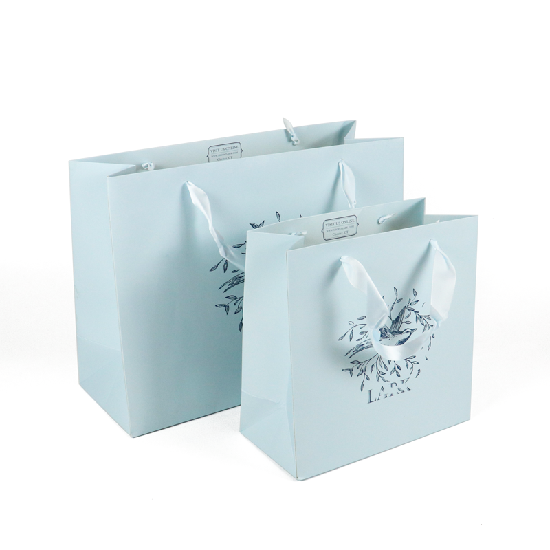 Two pastel blue paper shopping bags with white satin ribbon handles and delicate bird-and-branch illustration, perfect for luxury fashion or ethical lifestyle brands.