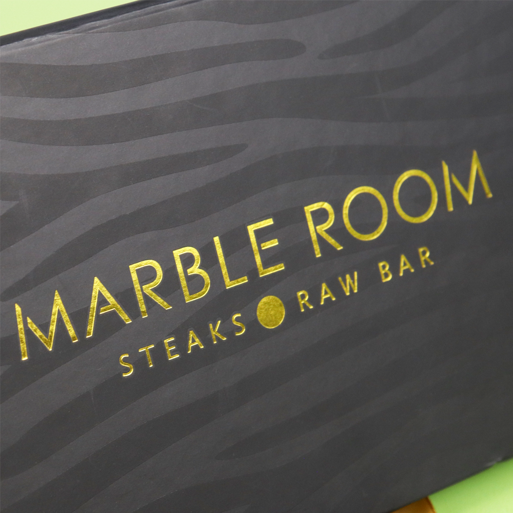 Close-up of gold foil-stamped "MARBLE ROOM STEAKS • RAW BAR" text on a black box with wavy pattern, showcasing elegant contrast and design focus.