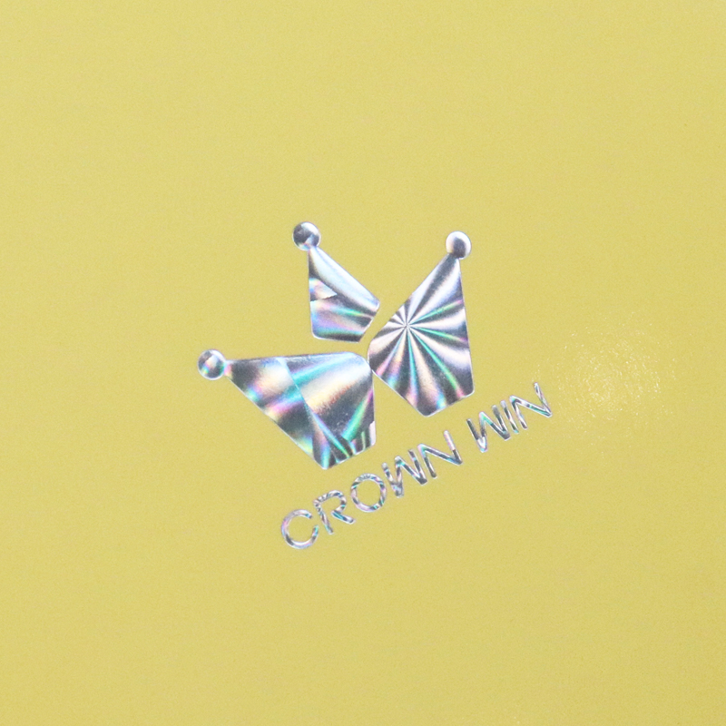 Close-up of a holographic foil-stamped logo "CROWN WIN" on a yellow surface, featuring iridescent crown design and reflective text.