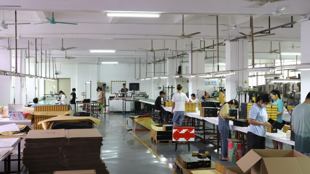 Wide view of a factory floor with workers assembling boxes at production lines, surrounded by stacks of cardboard, finished products, and machinery.