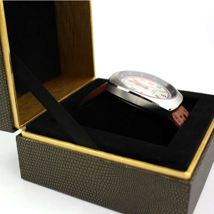 Open rigid box with black velvet interior and gold trim, holding a silver watch with brown leather strap secured by a black fabric strap.