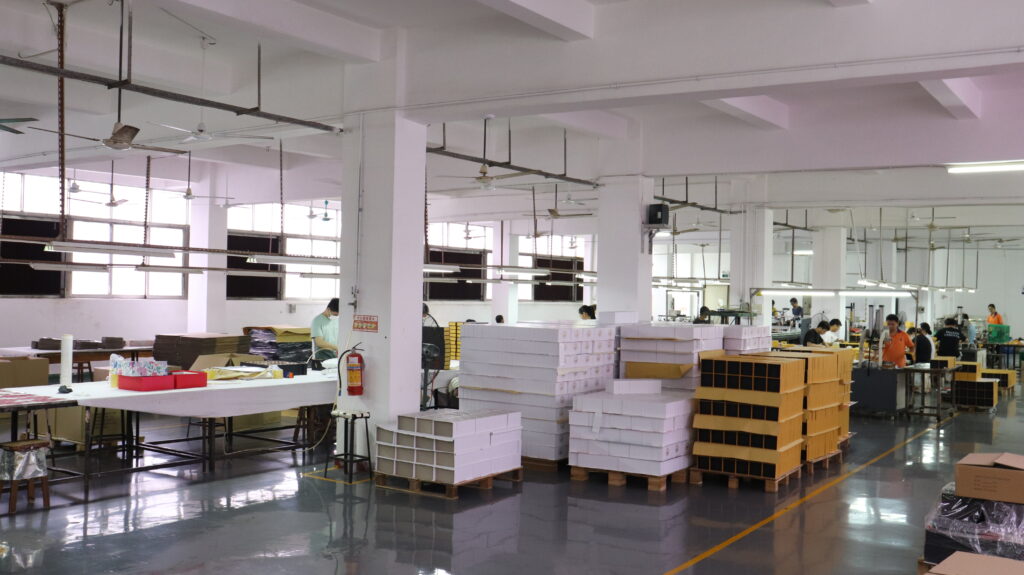 Factory production area with stacks of finished boxes and workers assembling packaging, illustrating the challenges of producing both disposable and cartridge packaging formats.