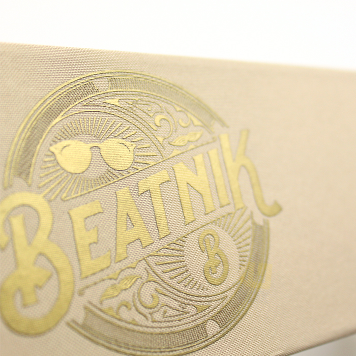 Close-up of gold foil-stamped "BEATNIK" logo with sunglasses motif on a beige textured paper surface, showcasing refined minimalism.