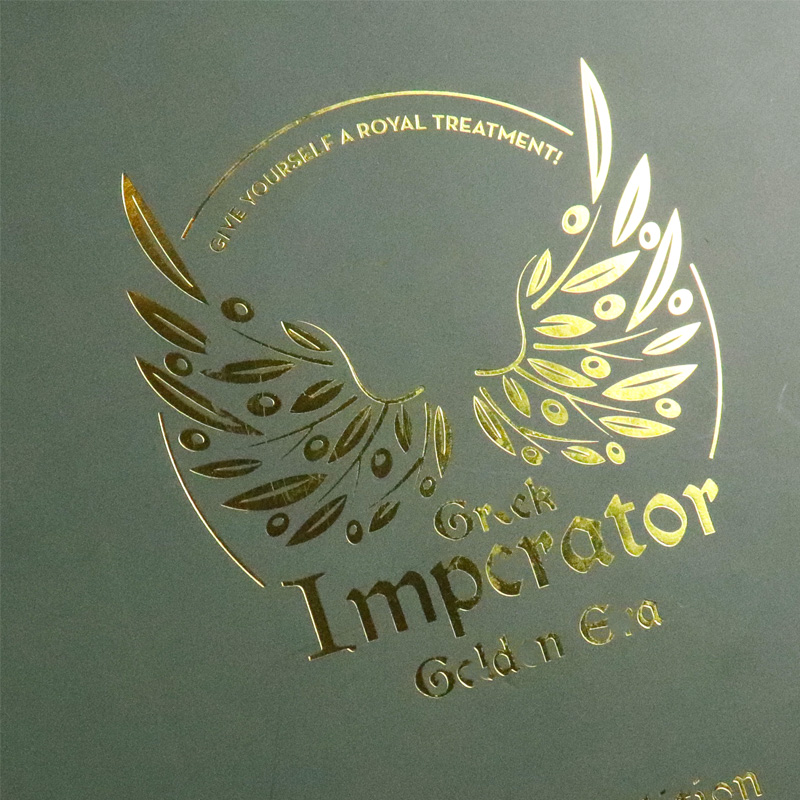 Close-up of gold foil-stamped logo featuring olive branches and text "Greek Imperator Golden Era" on a dark green matte surface.