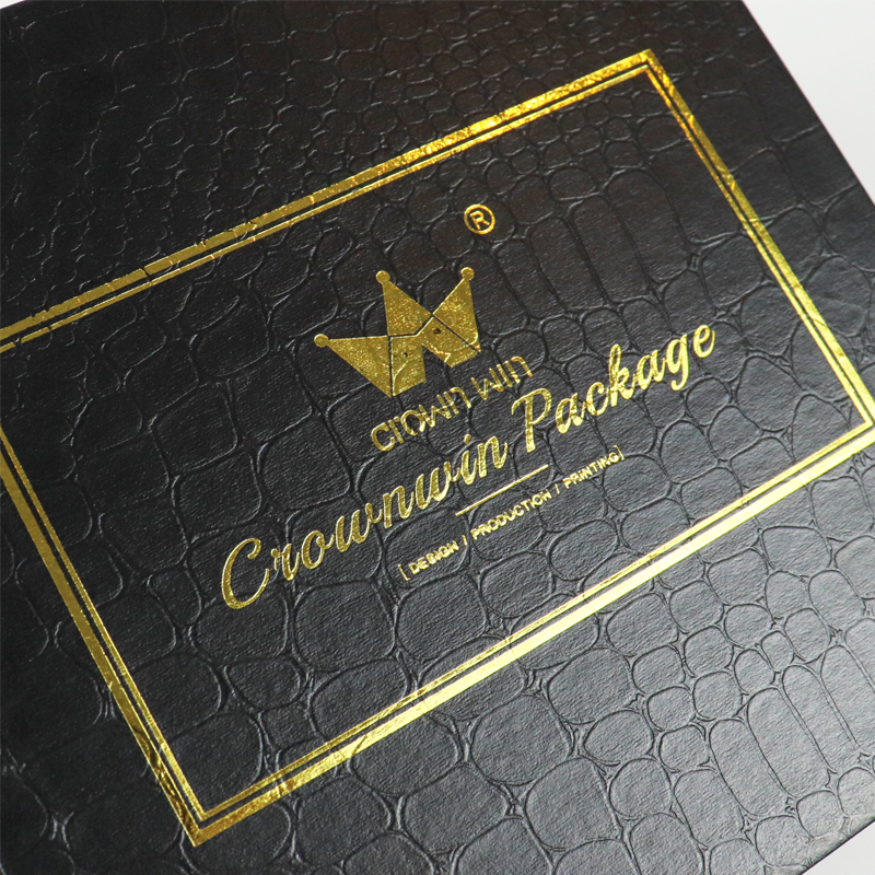 Close-up of a black rigid box with crocodile skin texture embossing and gold foil-stamped logo within a decorative border.