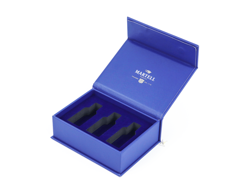 An open flip-top sapphire blue rigid cardboard box showcasing a metallic gold foil printed brand emblem and a matching blue velvet lined tray with multi-slot product compartments.